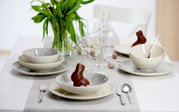 Elegantes 18-teiliges Geschirrset auf einem gedeckten Tisch mit Frühlingsdekoration, aus der Perspektive von vorne betrachtet.