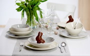 Elegantes 18-teiliges Geschirrset auf einem gedeckten Tisch mit Frühlingsdekoration, aus der Perspektive von vorne betrachtet.
