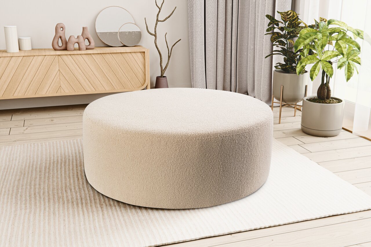 Runder, cremefarbener Pouf/Polsterhocker mit weichem Bouclé-Bezug im modernen Wohnzimmer auf hellem Teppich, Perspektive: leicht erhöhte Frontansicht.