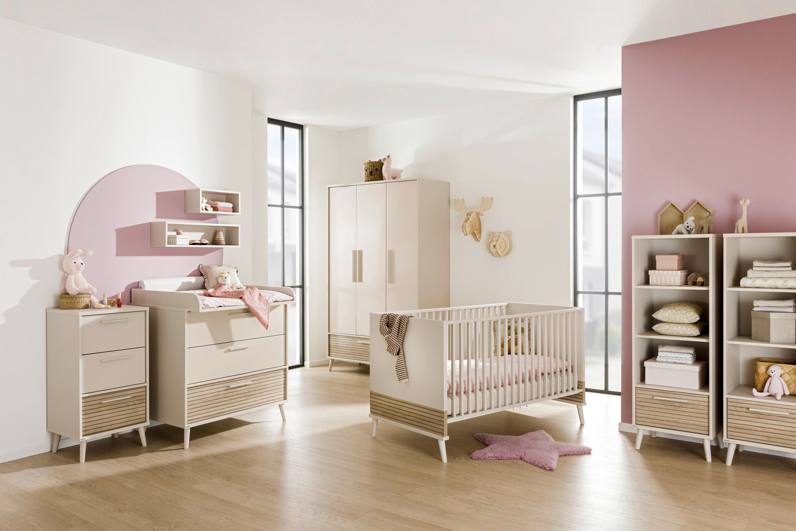Modernes Babyzimmer mit einem cremefarbenen Babybett, Wickelkommode und Schrank, dekoriert mit rosa Akzenten und Spielzeug, seitliche Perspektive.