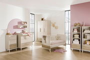 Modernes Babyzimmer mit einem cremefarbenen Babybett, Wickelkommode und Schrank, dekoriert mit rosa Akzenten und Spielzeug, seitliche Perspektive.