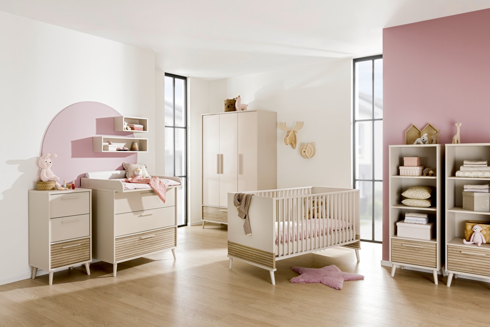 Moderne babykamer met een crèmekleurig babybedje, commode en kast, versierd met roze accenten en speelgoed, zijaanzicht.
