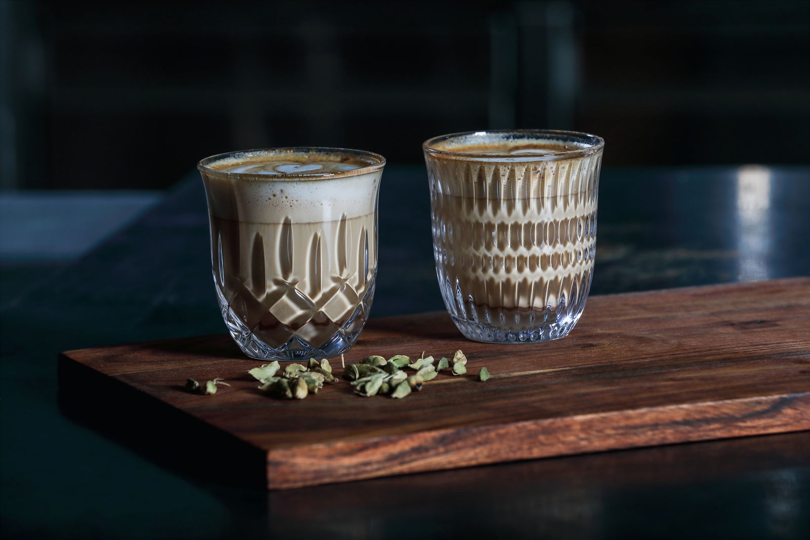 Zwei kunstvoll gestaltete Gläser mit Cappuccino und Flat White auf einem Holzbrett, umgeben von Kardamomkapseln, aus seitlicher Perspektive.