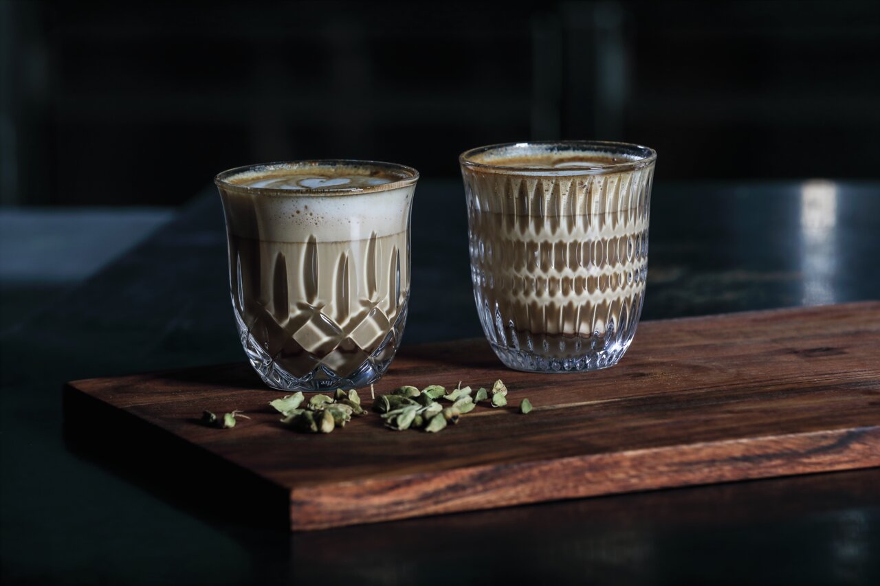 Twee kunstig vormgegeven glazen met cappuccino en flat white op een houten plank, omringd door kardemompeulen, gezien vanuit een zijaanzicht.