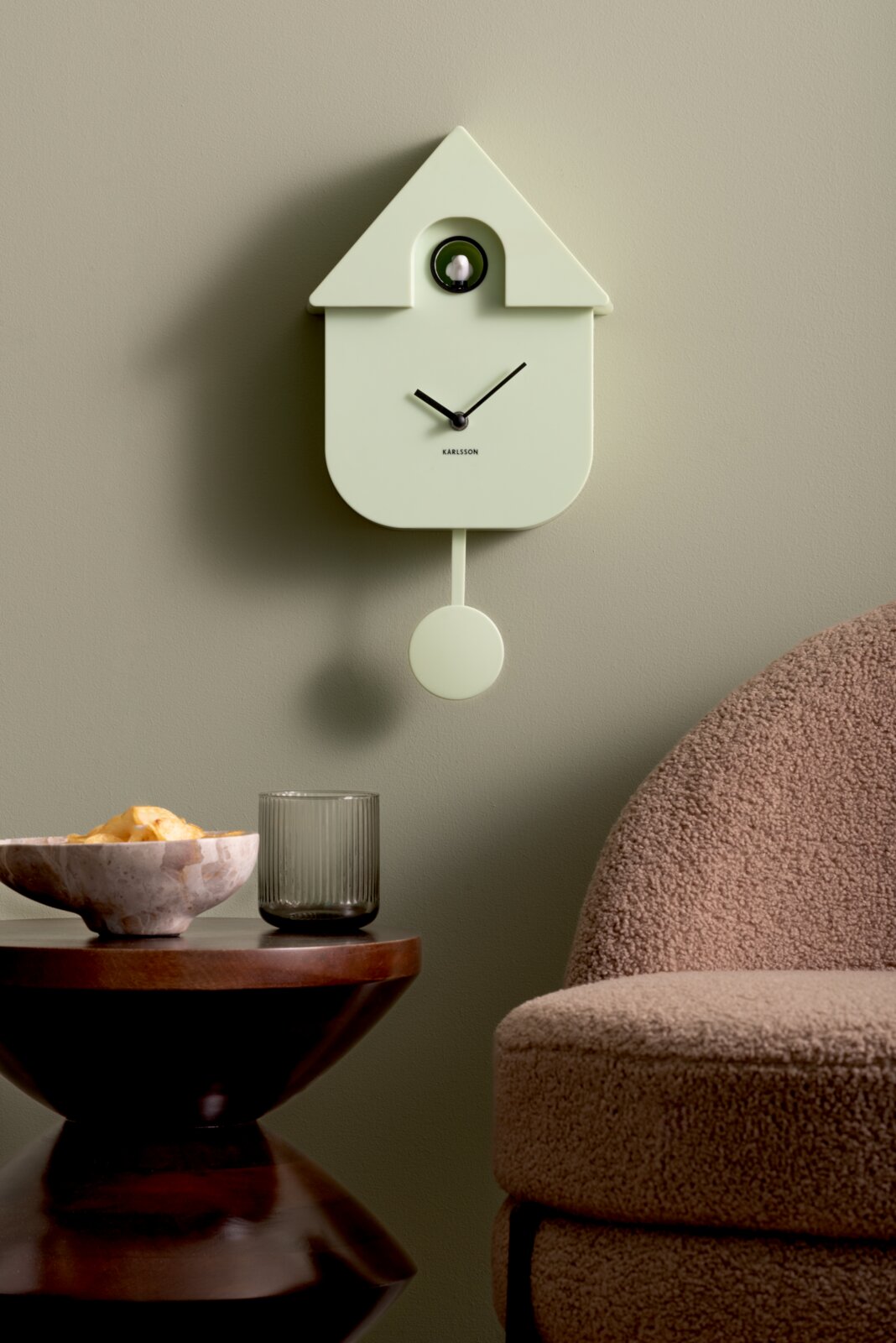 Moderne wandklok in de vorm van een huisje met slinger, in een lichte groene tint, hangend aan een groene muur. Vooraanzicht.