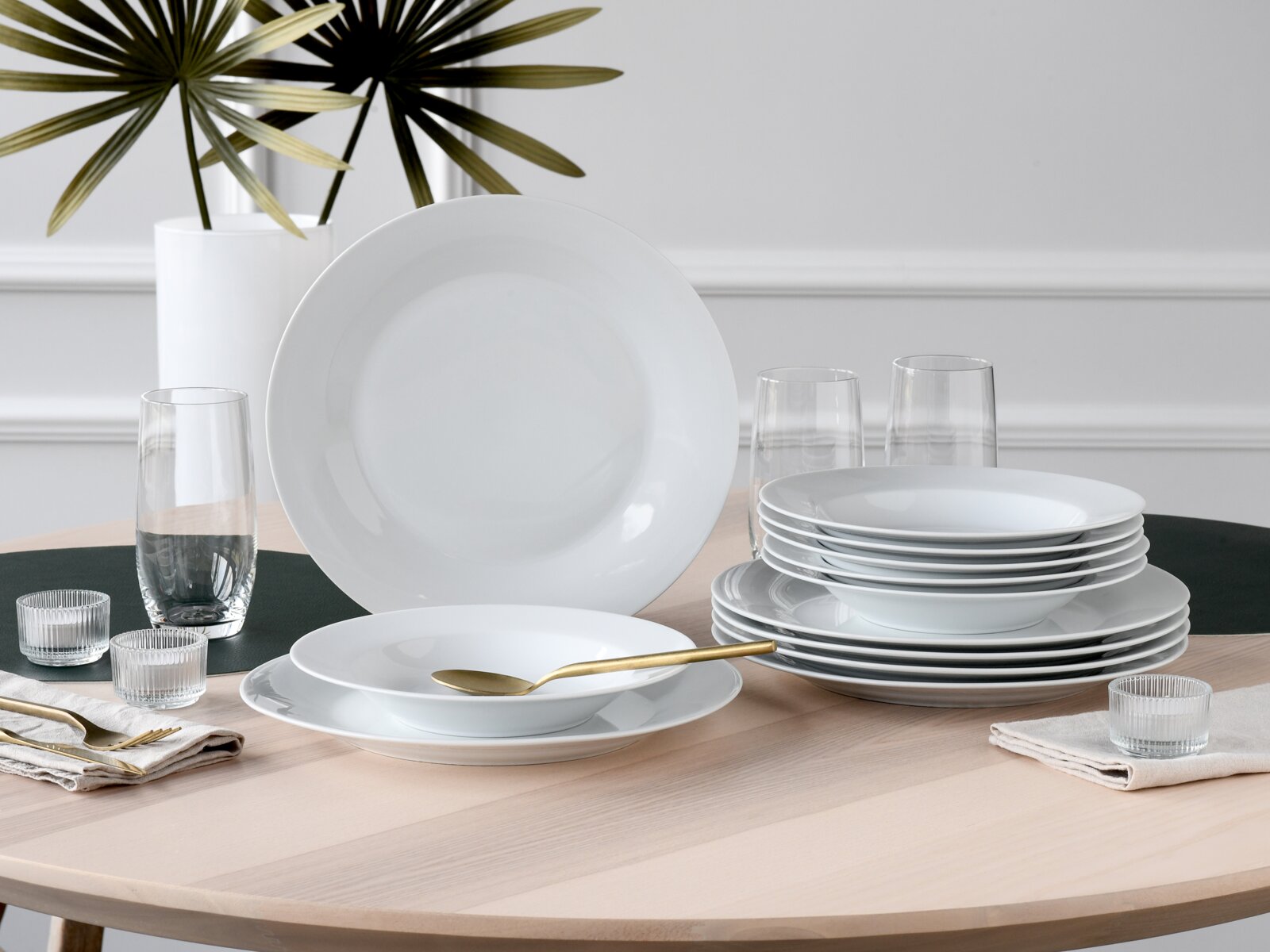 Weißes, minimalistisches Geschirrset mit flachen und tiefen Tellern auf einem hellen Holztisch, seitliche Frontperspektive mit leicht erhöhter Ansicht; dekoriert mit Glasbechern, goldfarbenem Besteck und Servietten.