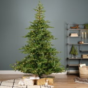 Vooraanzicht van een volle groene kunstkerstboom met warmwitte verlichting, sfeervol opgesteld in een modern interieur met cadeaus eronder