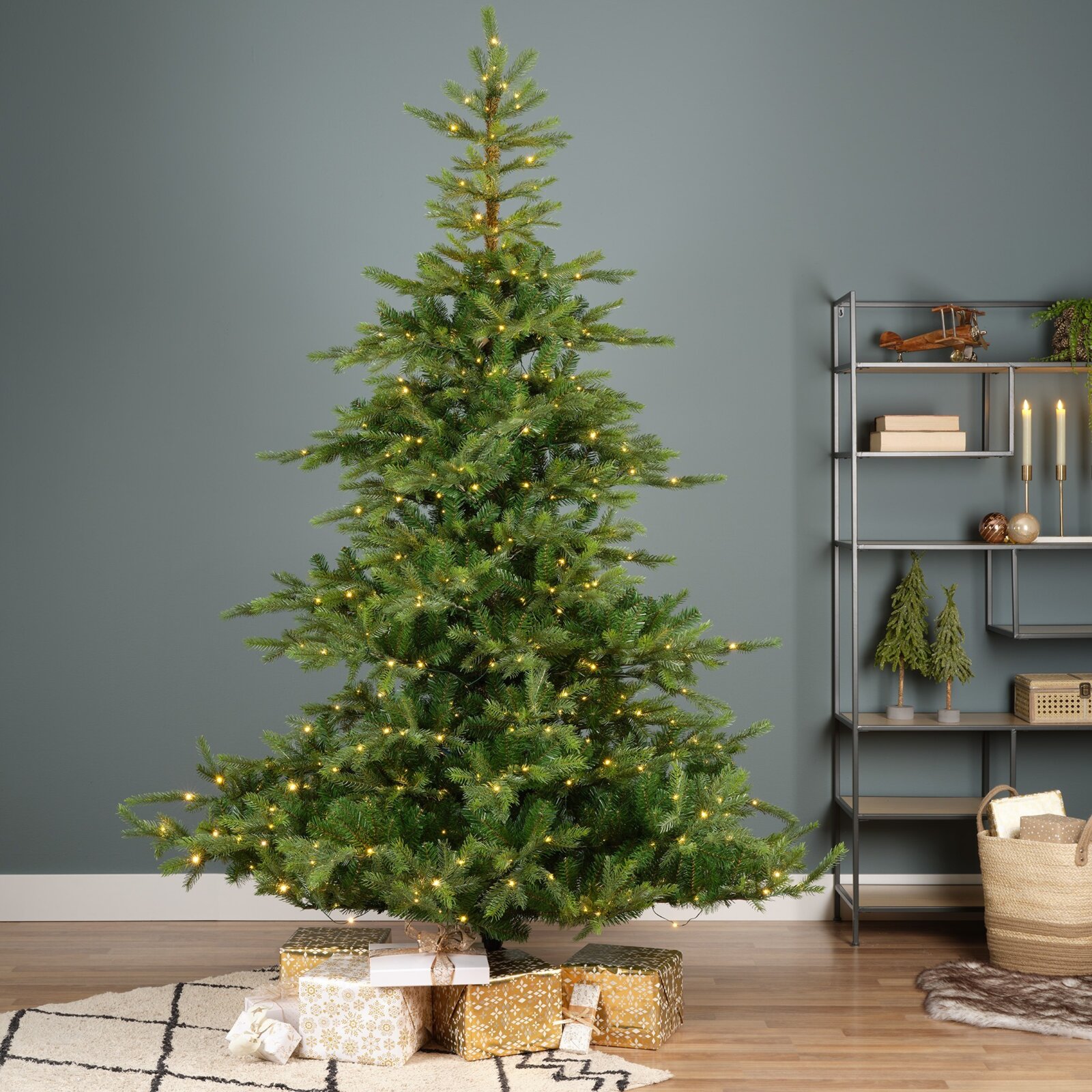 Vooraanzicht van een volle groene kunstkerstboom met warmwitte verlichting, sfeervol opgesteld in een modern interieur met cadeaus eronder