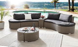 Moderne Lounge-Sitzgruppe aus Rattan mit grauen Kissen, bestehend aus einem geschwungenen Sofa und einem runden Couchtisch, auf einer Terrasse mit Blick auf einen Pool und Palmen. Perspektive von vorne.