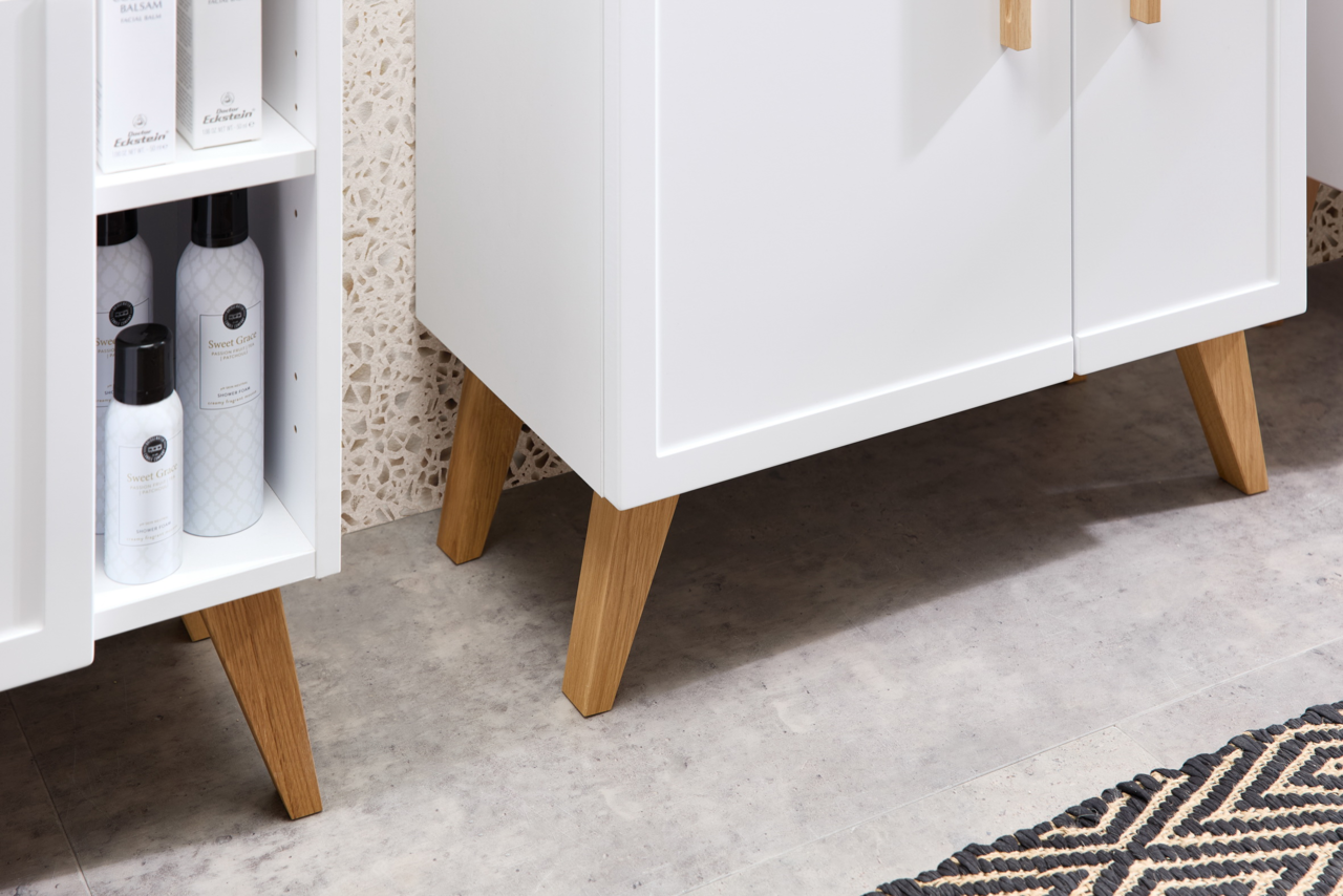 Nahaufnahme eines weißen Schranks mit Holzfüßen auf einem Fliesenboden, seitliche Perspektive.