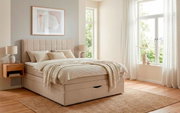 Beiges, gepolstertes Boxspringbett mit gestepptem Kopfteil und integriertem Bettkasten in hellem Schlafzimmer, fotografiert in frontaler Perspektive mit leichtem Blick von rechts.