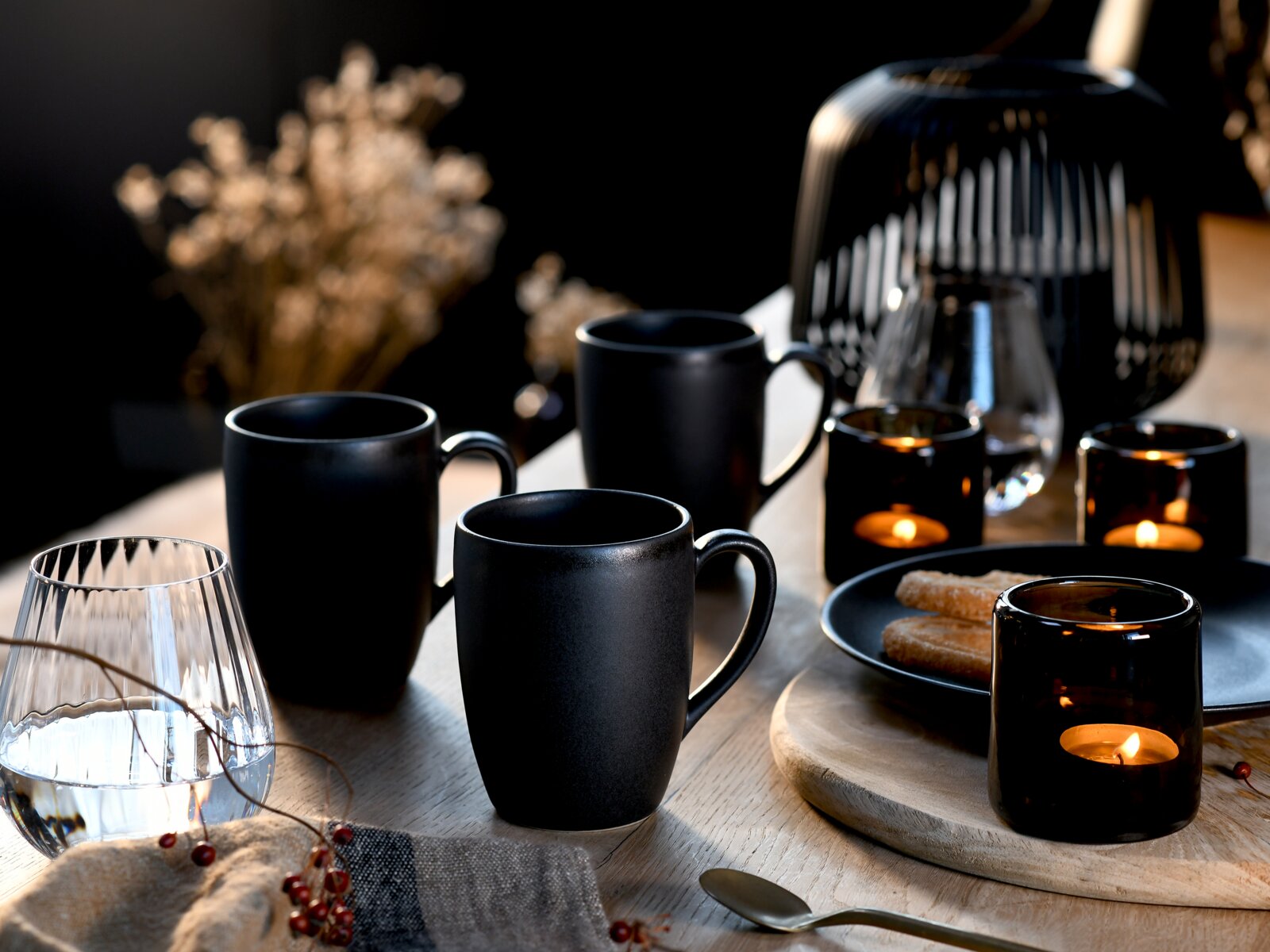 Drei matte schwarze Keramikbecher mit Henkel auf einem gedeckten Holztisch mit Kerzenlicht und Gebäck, schräg von vorn aus Tischhöhe fotografiert.