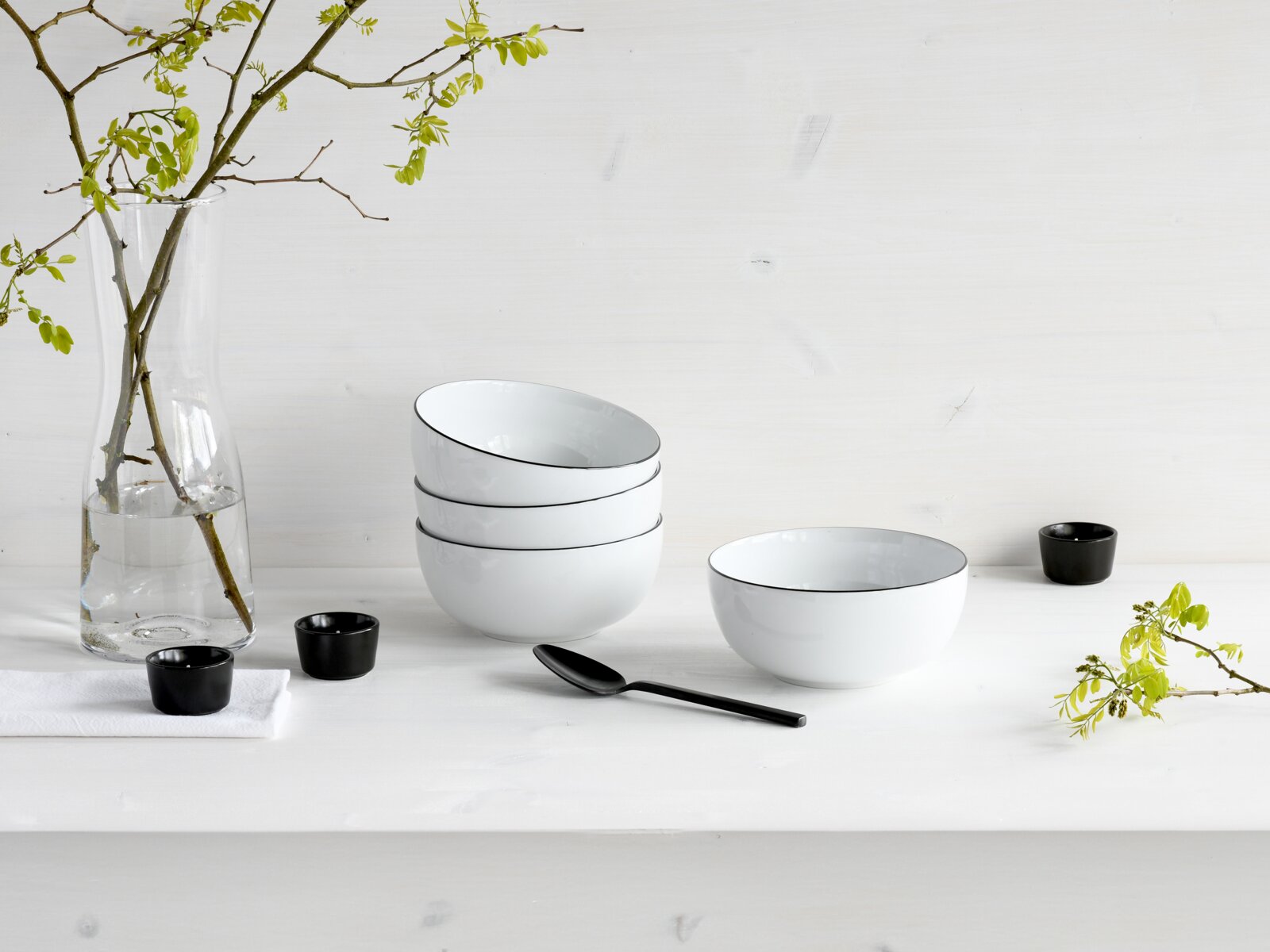 Minimalistisches Stillleben: vier weiße Schalen mit schwarzem Rand (eine seitlich aufliegend), daneben schwarze Löffel und kleine Schälchen sowie Glasvase mit Zweigen auf weißem Tisch, fotografiert in leichter Seitenansicht auf Augenhöhe.