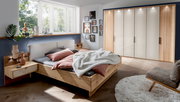 Modernes Futonbett aus Holz in einem stilvoll eingerichteten Schlafzimmer mit Blick auf das Bett von der Seite. Das Bett ist mit rosa und grauen Kissen sowie einer weißen Decke dekoriert. Im Hintergrund sind ein großer Kleiderschrank und ein Fenster mit v