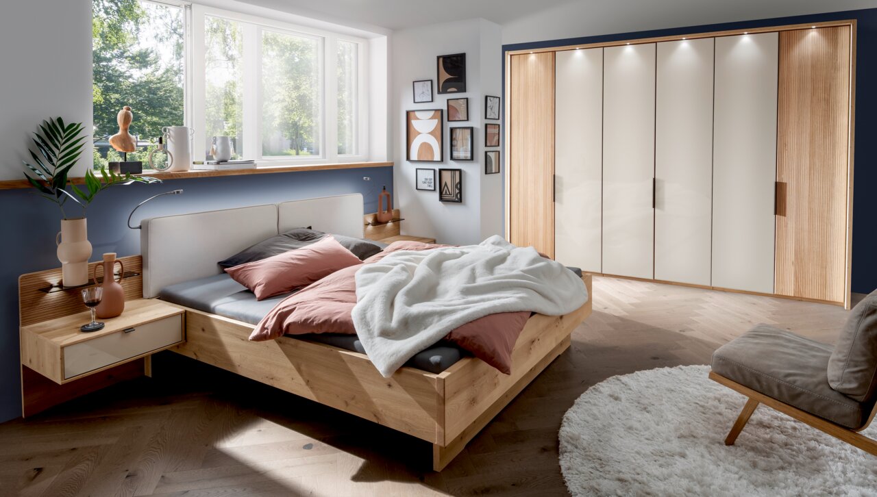 Modernes Futonbett aus Holz in einem stilvoll eingerichteten Schlafzimmer mit Blick auf das Bett von der Seite. Das Bett ist mit rosa und grauen Kissen sowie einer weißen Decke dekoriert. Im Hintergrund sind ein großer Kleiderschrank und ein Fenster mit v