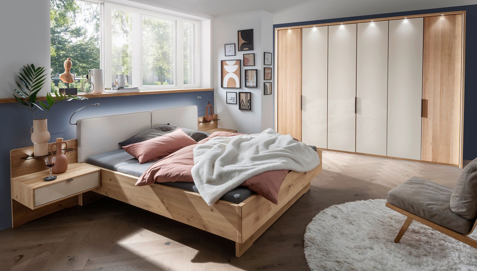 Modernes Futonbett aus Holz in einem stilvoll eingerichteten Schlafzimmer mit Blick auf das Bett von der Seite. Das Bett ist mit rosa und grauen Kissen sowie einer weißen Decke dekoriert. Im Hintergrund sind ein großer Kleiderschrank und ein Fenster mit v