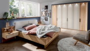 Modernes Futonbett aus Holz in einem stilvoll eingerichteten Schlafzimmer mit Blick auf das Bett von der Seite. Das Bett ist mit rosa und grauen Kissen sowie einer weißen Decke dekoriert. Im Hintergrund sind ein großer Kleiderschrank und ein Fenster mit v
