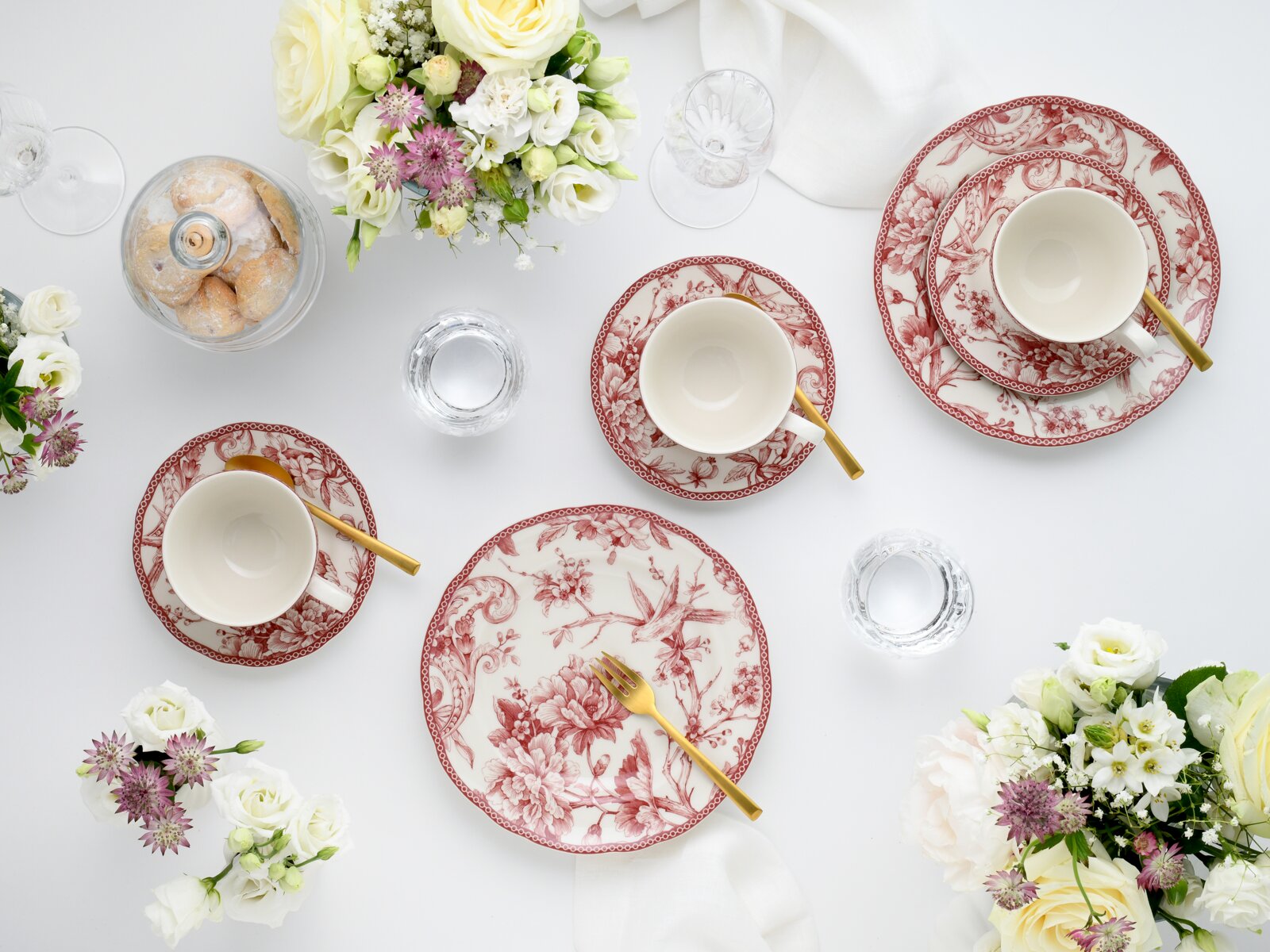 Elegantes Tafelservice mit rot-weiß gemusterten Tellern und Tassen sowie goldfarbenem Besteck auf weißer Tischdecke, dekoriert mit Blumen und Gebäck, fotografiert aus der Vogelperspektive (Top-Down).