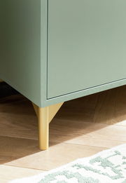 Nahaufnahme eines grünen Waschbeckenunterschranks mit goldenen Beinen auf einem Holzboden, aus seitlicher Perspektive.