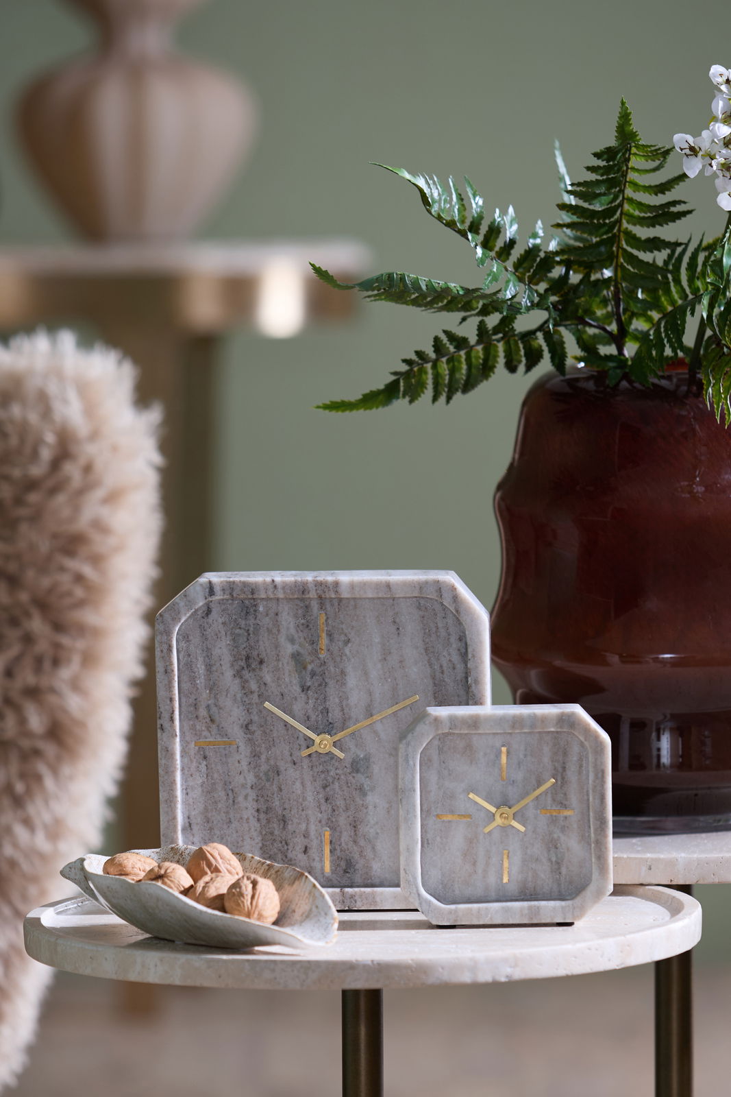 Light&Living Uhr PABLIA Zwei stilvolle Tischuhren aus Marmor mit goldenen Zeigern, auf einem runden Tisch arrangiert, im Hintergrund eine Pflanze und ein dekorativer Topf. Perspektive: Frontalansicht.