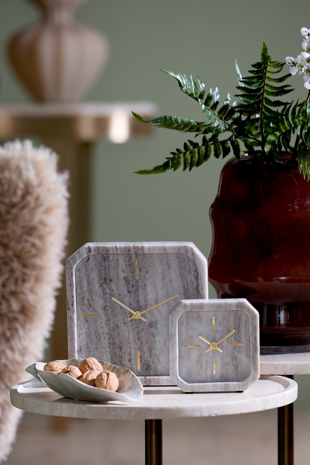 Light&Living Uhr PABLIA Zwei stilvolle Tischuhren aus Marmor mit goldenen Zeigern, auf einem runden Tisch arrangiert, im Hintergrund eine Pflanze und ein dekorativer Topf. Perspektive: Frontalansicht.
