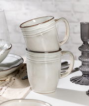 Zwei gestapelte, cremefarbene Keramiktassen mit dezentem Rillenmuster, seitliche Perspektive, auf einem gedeckten Tisch mit weiteren Geschirrteilen.