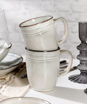 Zwei gestapelte, cremefarbene Keramiktassen mit dezentem Rillenmuster, seitliche Perspektive, auf einem gedeckten Tisch mit weiteren Geschirrteilen.