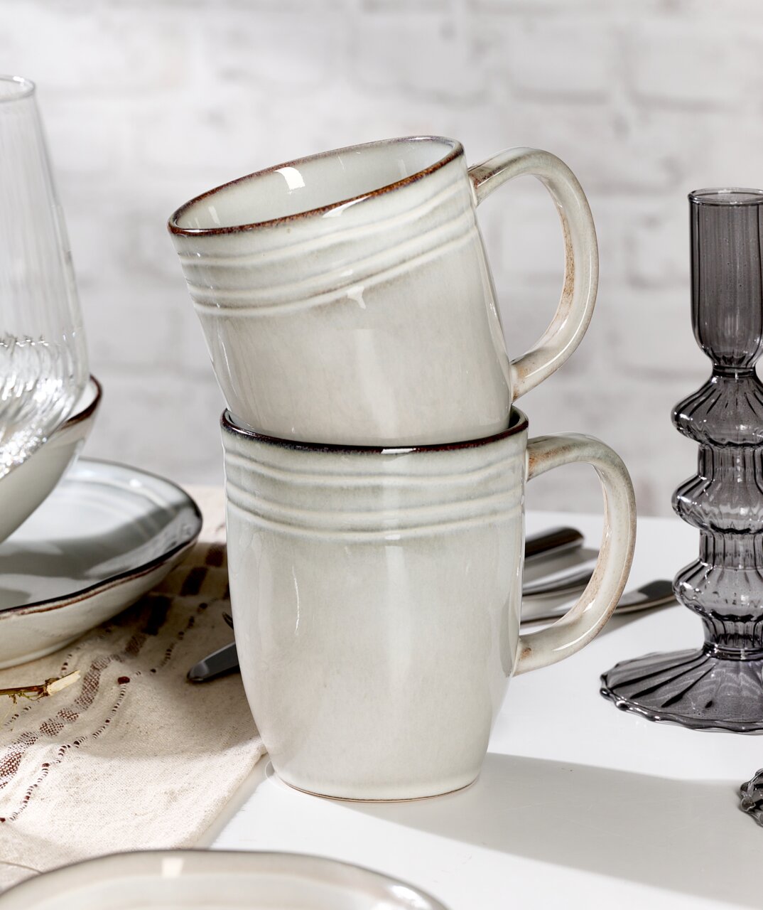 Zwei gestapelte, cremefarbene Keramiktassen mit dezentem Rillenmuster, seitliche Perspektive, auf einem gedeckten Tisch mit weiteren Geschirrteilen.