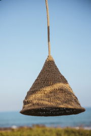 Outdoor-Hängeleuchte aus geflochtenem Material in Kegelform, vor blauem Himmel und Meereshintergrund, seitliche Perspektive