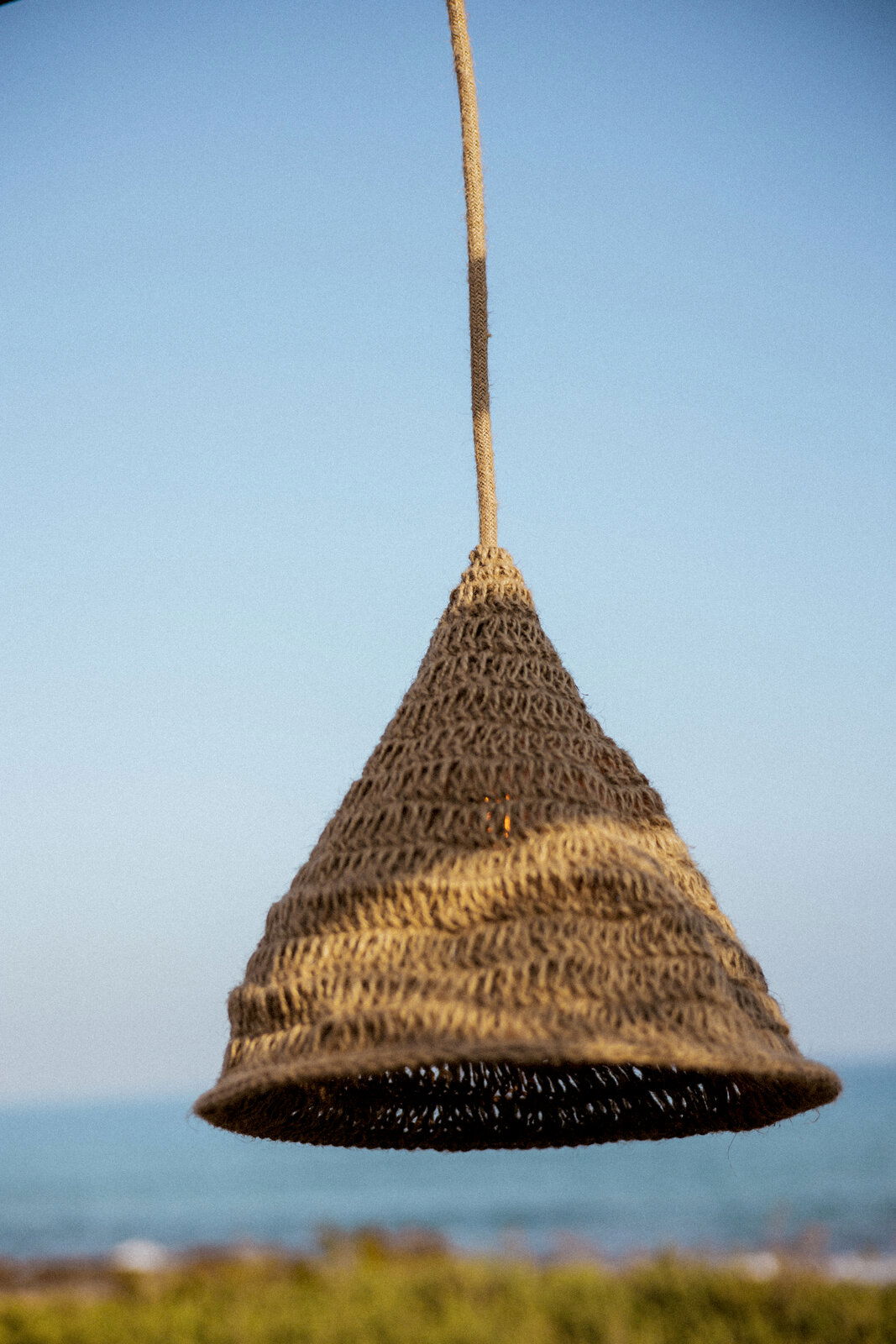 Outdoor-Hängeleuchte aus geflochtenem Material in Kegelform, vor blauem Himmel und Meereshintergrund, seitliche Perspektive