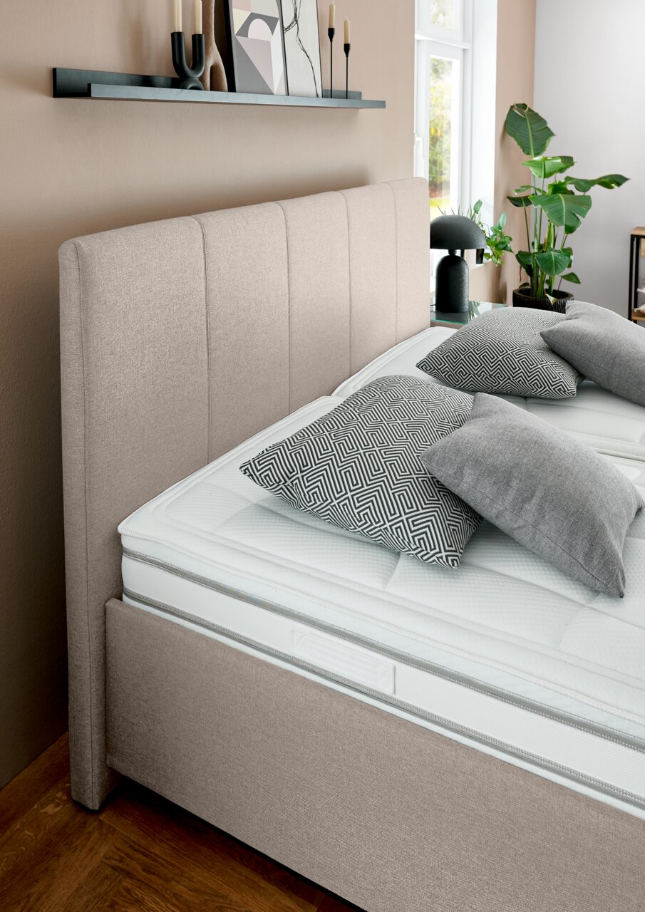 Beige gestoffeerd bed met hoog hoofdeinde, gezien vanaf de zijkant, versierd met grijze en gedessineerde kussens, in een moderne slaapkamer.