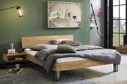 Modernes Holzbett mit schlichtem Kopfteil und Metallfüßen, grau bezogen und mit Kissen dekoriert, im Schlafzimmer-Setting; Perspektive: frontale Seitenansicht leicht von rechts.
