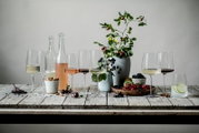 Stillleben in Frontansicht auf rustikalem Holztisch mit mehreren Weingläsern (Weiß-, Rosé- und Rotwein), Glasflaschen, Beeren und Nüssen sowie einer Vase mit Zweigen vor grauem Hintergrund