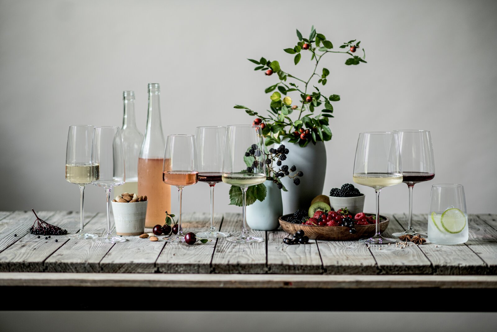 Stillleben in Frontansicht auf rustikalem Holztisch mit mehreren Weingläsern (Weiß-, Rosé- und Rotwein), Glasflaschen, Beeren und Nüssen sowie einer Vase mit Zweigen vor grauem Hintergrund