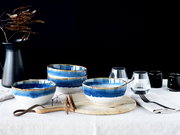 Blau-weiß glasierte Keramikschalen im Set auf gedecktem Tisch mit Holzbrett, Gläsern und Besteck vor schwarzem Hintergrund, Perspektive: frontale Tischhöhe leicht seitlich