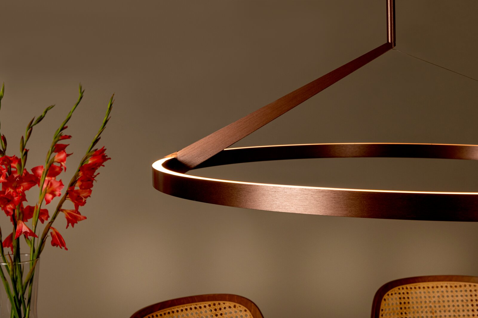 Moderne ringförmige LED-Pendelleuchte in Bronze/Kupferoptik mit warmweißem Licht, seitliche Perspektive in Wohnraum-Szene mit roten Blumen und Stühlen im Hintergrund