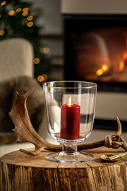 Windlicht mit roter Kerze auf einem rustikalen Holztisch, im Hintergrund ein gemütliches Kaminfeuer, seitliche Perspektive.