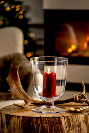 Windlicht mit roter Kerze auf einem rustikalen Holztisch, im Hintergrund ein gemütliches Kaminfeuer, seitliche Perspektive.