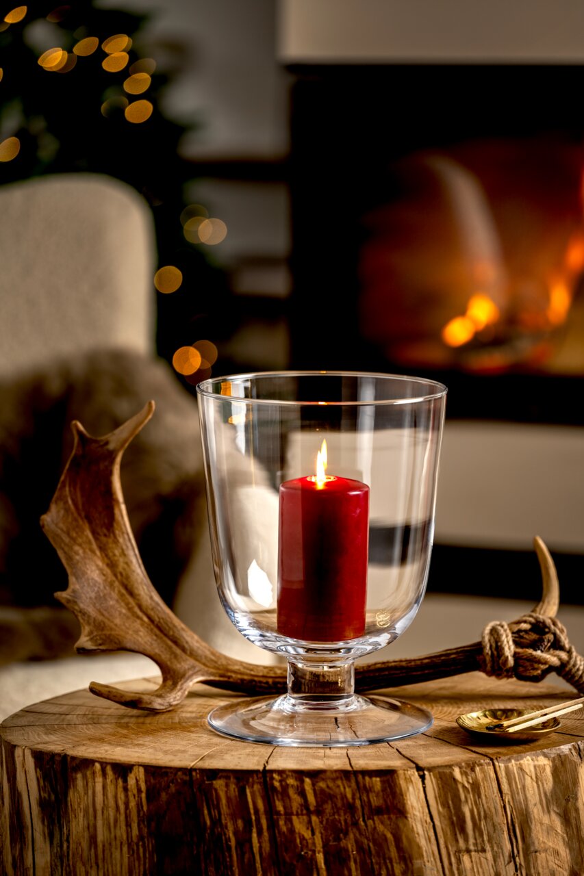 Windlicht mit roter Kerze auf einem rustikalen Holztisch, im Hintergrund ein gemütliches Kaminfeuer, seitliche Perspektive.