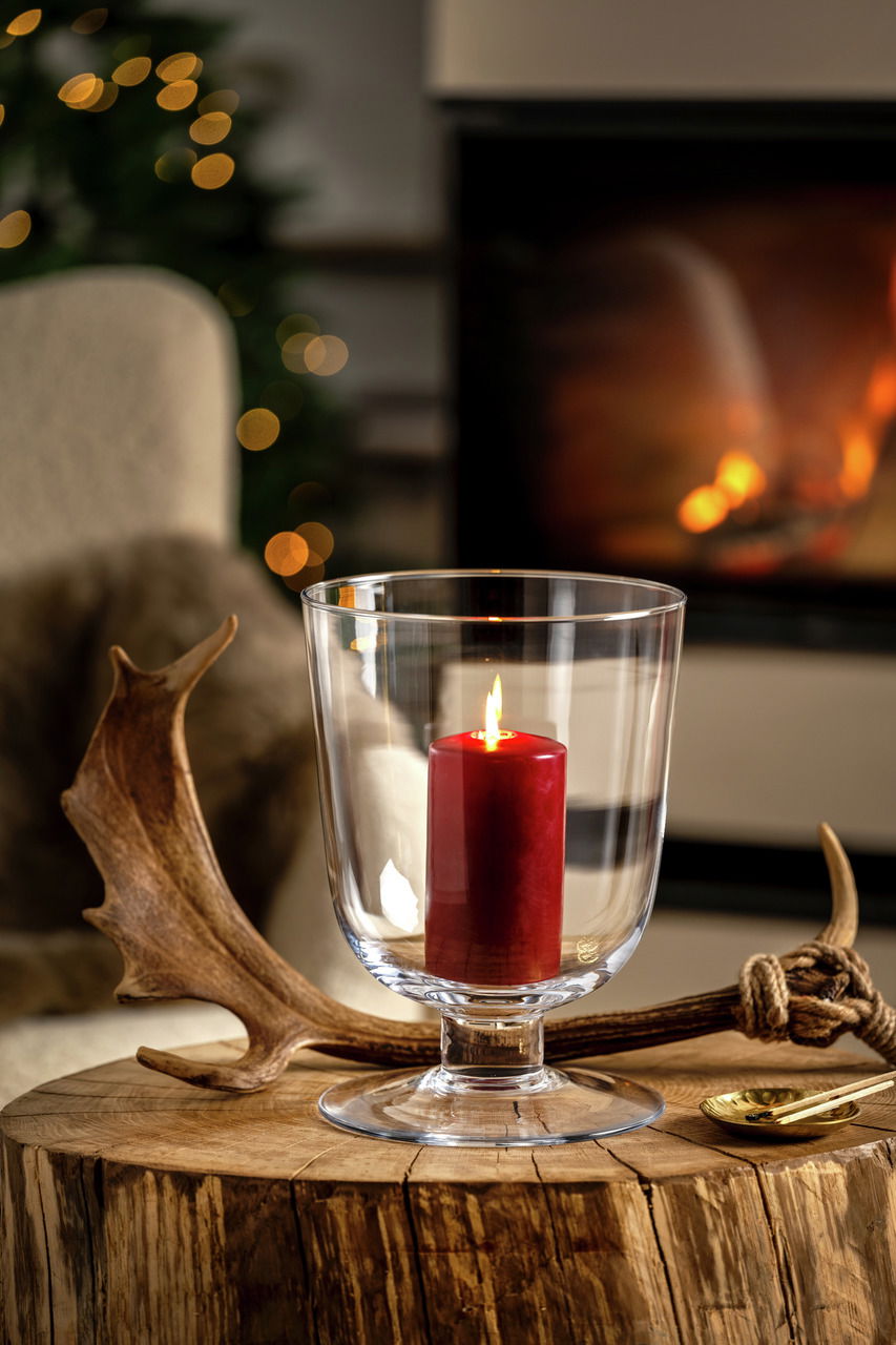 Windlicht mit roter Kerze auf einem rustikalen Holztisch, im Hintergrund ein gemütliches Kaminfeuer, seitliche Perspektive.