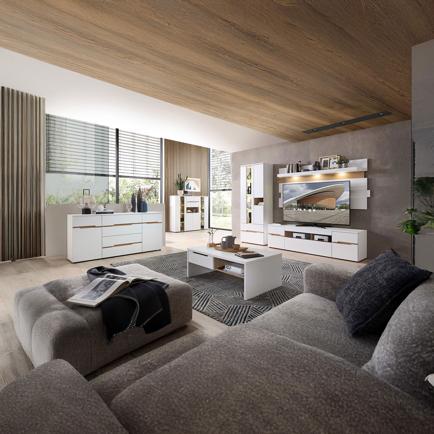 Modernes Wohnzimmer mit weißem Möbelset, großer grauer Couch, Holzdecke, TV-Wand und viel Tageslicht durch große Fenster.