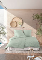Bettwäsche-Set in sanftem Grün auf einem Bett in einem modern eingerichteten Raum mit Pflanzen und dekorativen Körben an der Wand, aus der Perspektive von vorne betrachtet.