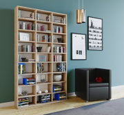 Großes, helles Holz-Bücherregal mit vielen Fächern, gefüllt mit Büchern und Deko, in modernem Wohnzimmer neben schwarzem Sessel; Ansicht schräg von vorn.