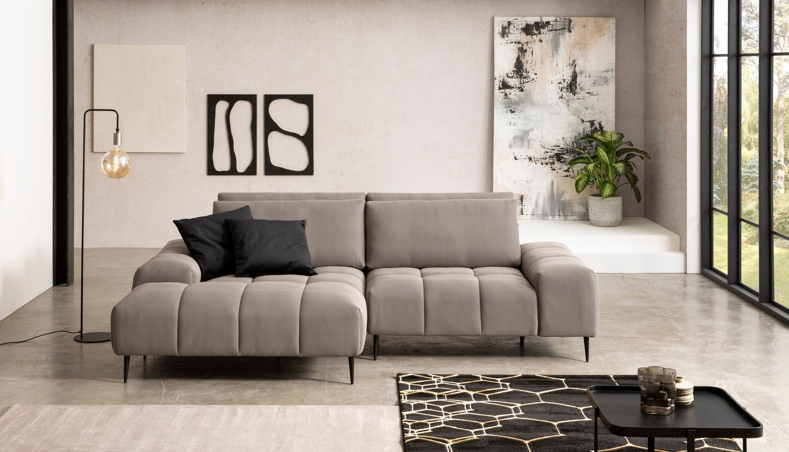 Moderne Longchairkombination in einem stilvollen Wohnzimmer, frontal betrachtet. Die Couch ist in einem hellen Grauton gehalten und mit schwarzen Kissen dekoriert. Im Hintergrund sind Kunstwerke und eine Pflanze zu sehen.