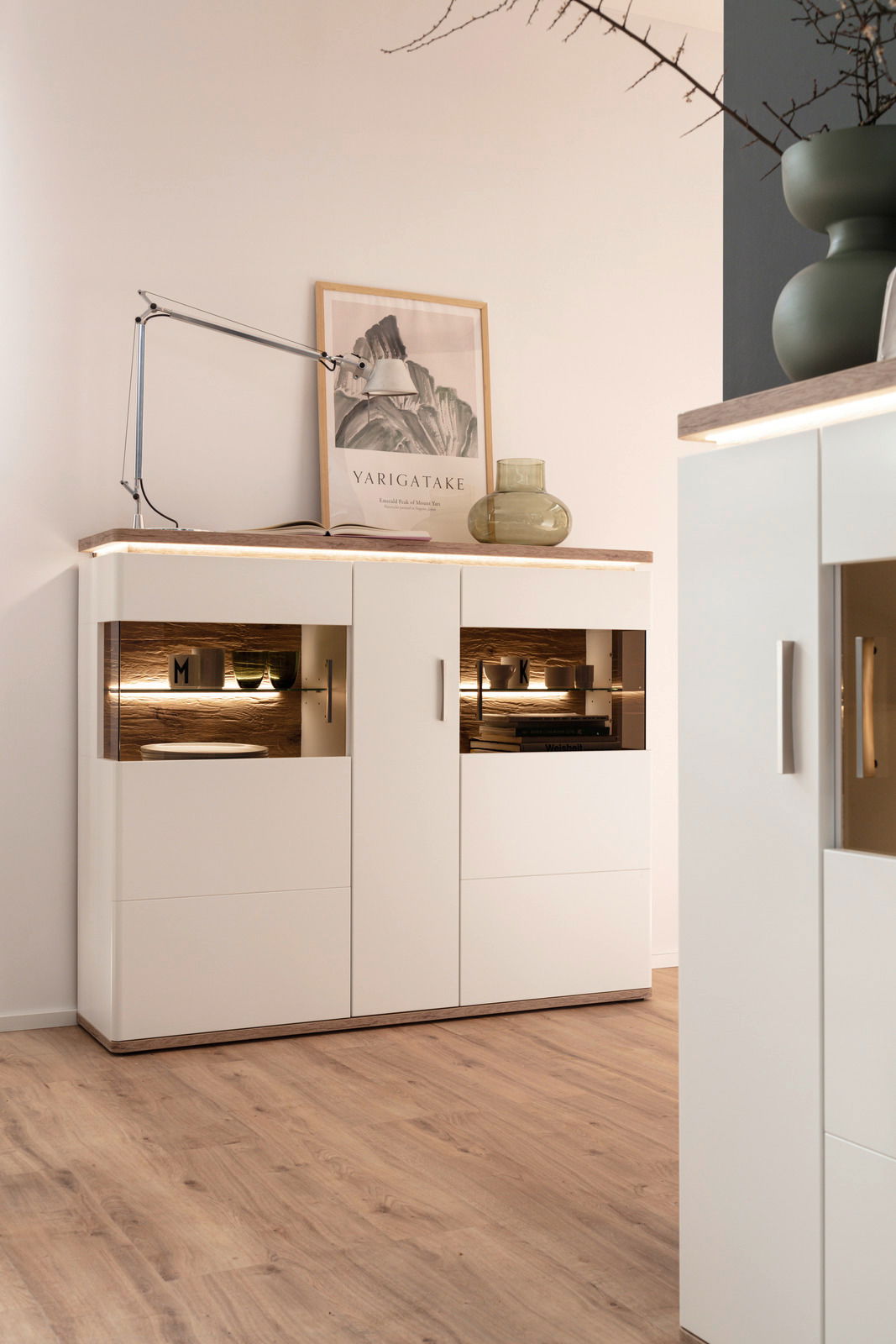 Modernes Highboard in Weiß mit Holzdetails, dekoriert mit einer Lampe und einem Glasgefäß, seitliche Perspektive
