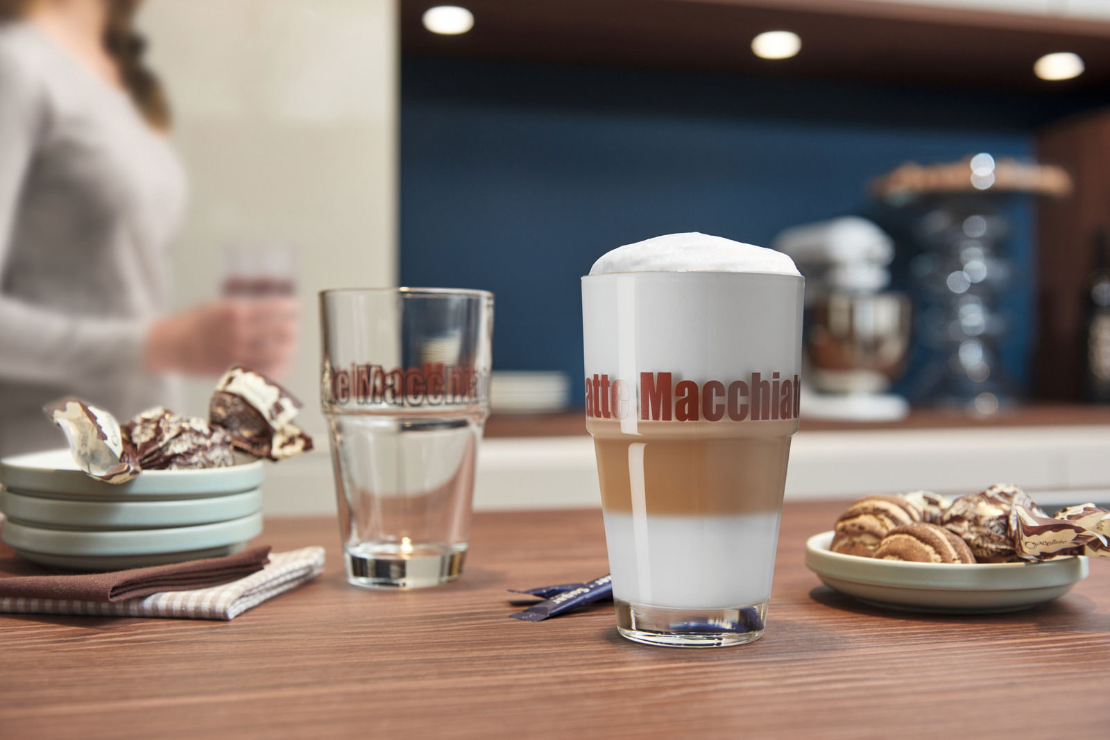 Latte Macchiato Becher Solo auf einem Tisch in einer modernen Küche, gefüllt mit einem geschichteten Latte Macchiato, im Vordergrund aus seitlicher Perspektive.