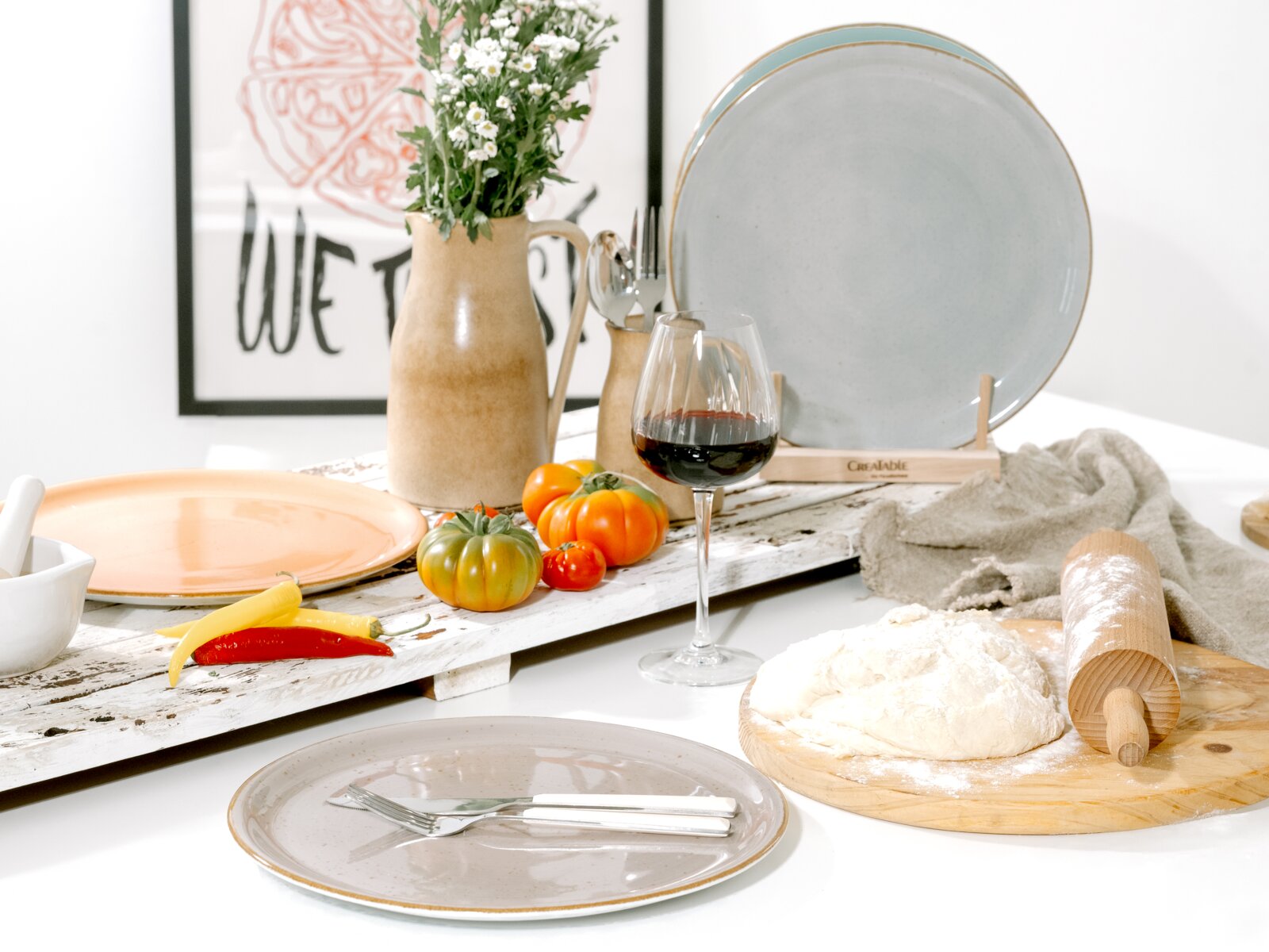 Rustikales Tisch- und Küchen-Stillleben mit Keramikgeschirr (grauer Teller und großer grauer Servierteller im Ständer), Krug mit Blumen, Rotweinglas, Tomaten, Chilischoten sowie Teig und Nudelholz auf Holzbrett, aus leicht schräger Frontperspektive aufgen