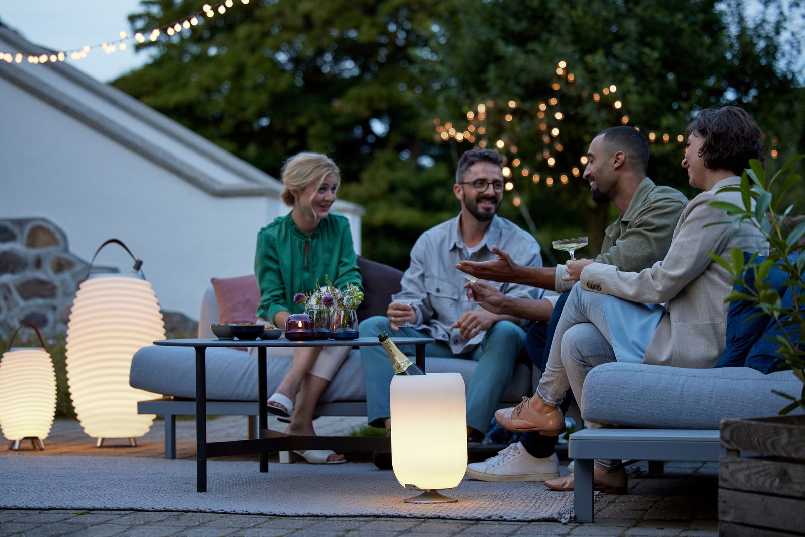 Eine Gruppe von vier Personen sitzt entspannt auf einer Terrasse bei Dämmerung. Im Vordergrund steht eine leuchtende Akku-Leuchte, die auch als Lautsprecher dient. Die Szene ist von einer gemütlichen Atmosphäre mit Lichterketten im Hintergrund geprägt.