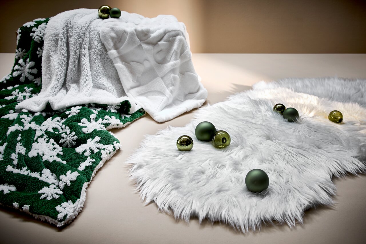 Twee knuffeldekens in kerststijl, een groene met witte sneeuwvlokken en een witte met een subtiel patroon, versierd met groene kerstballen, gefotografeerd vanuit een zijaanzicht.