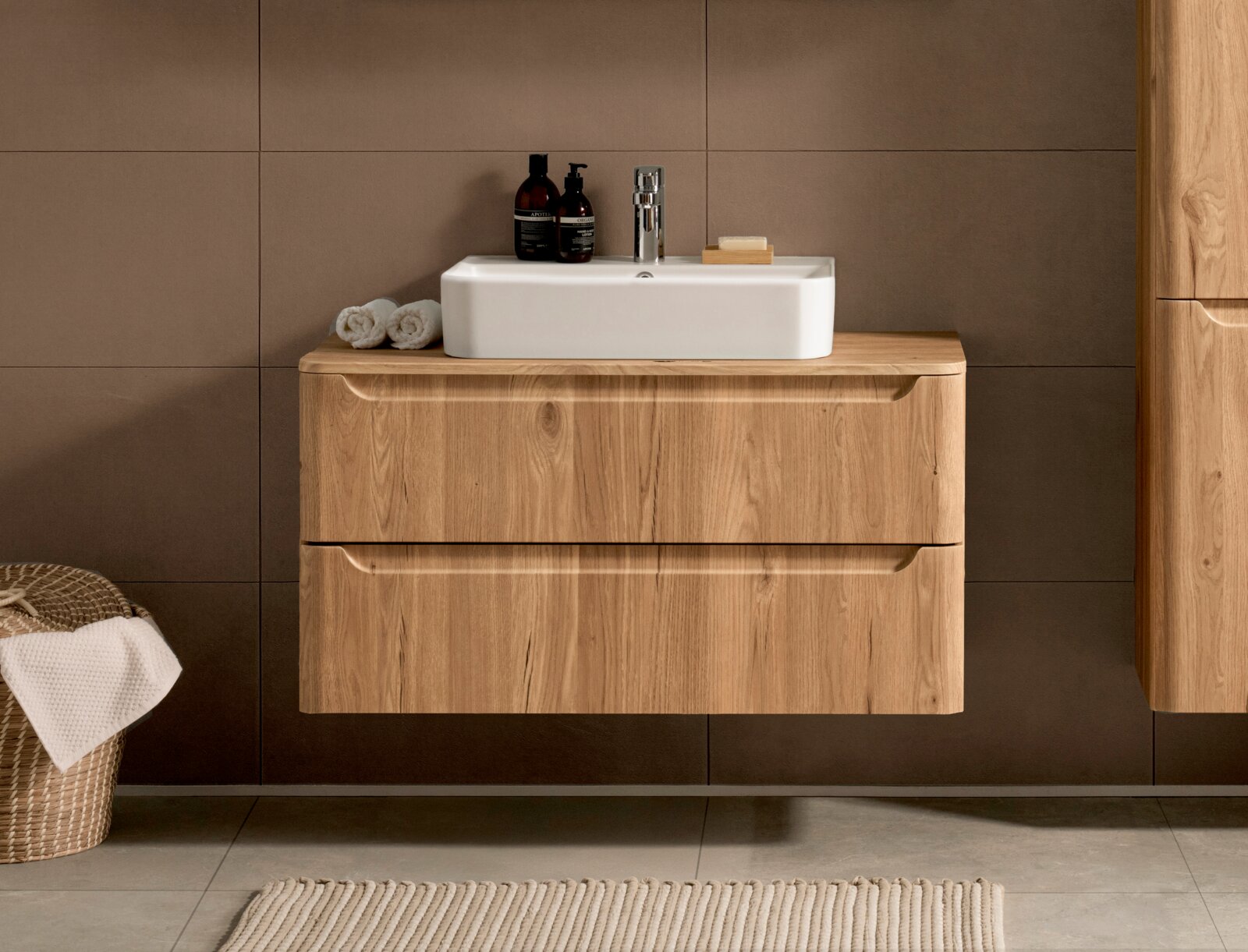 Frontansicht eines wandhängenden Waschtischunterschranks aus hellem Holz mit zwei Schubladen und weißem Aufsatzwaschbecken, chromfarbenem Wasserhahn und Bad-Accessoires vor brauner Fliesenwand
